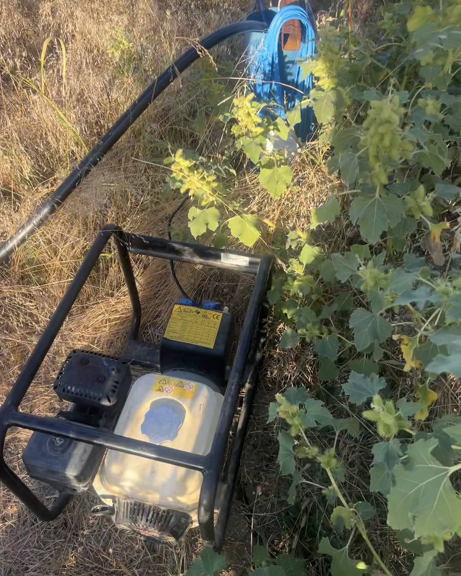 Motopompă pe benzină lăsată în iarbă la marginea unui teren de irigat, cu furtunul de aspirație și colacul de furtun plat alături